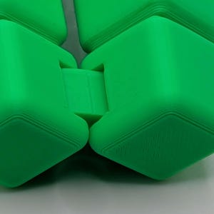 May include: Close-up of a bright green, articulated cube toy. The toy is made of several connected cube-shaped pieces. The edges of the cubes have a ridged design. The toy is on a white surface.