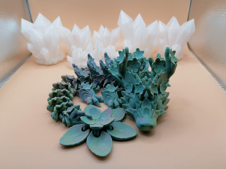 May include: A teal and green articulated dragon figurine with a flower and white crystal formations. The dragon has a detailed, textured design. The flower has six petals. The crystals are clustered together.