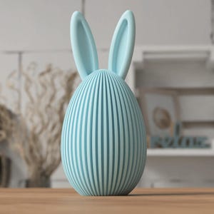 Puede incluir: Un objeto decorativo en forma de huevo azul claro con l&iacute;neas verticales y orejas de conejo. La decoraci&oacute;n con tem&aacute;tica de Pascua est&aacute; hecha de un material s&oacute;lido y descansa sobre una superficie de madera. El fondo incluye una estanter&iacute;a borrosa y plantas secas.
