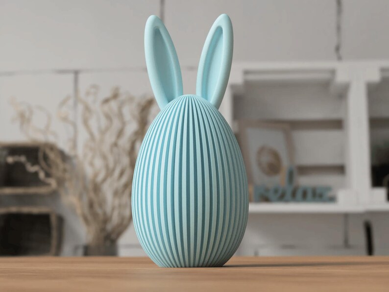 Puede incluir: Un objeto decorativo en forma de huevo azul claro con l&iacute;neas verticales y orejas de conejo. La decoraci&oacute;n con tem&aacute;tica de Pascua est&aacute; hecha de un material s&oacute;lido y descansa sobre una superficie de madera. El fondo incluye una estanter&iacute;a borrosa y plantas secas.