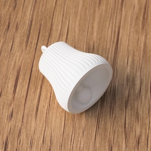 May include: A white, bell-shaped decorative object with a textured, ridged surface. The object has a small loop at the top and a smooth interior. The bell is resting on a wooden surface.