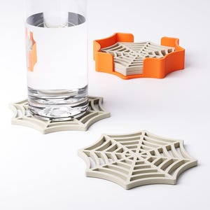 May include: A set of spiderweb-shaped coasters in light gray, with an orange holder. A tall glass of water sits on one coaster. The coasters are made of a durable material and are designed to protect surfaces from water rings. The set is ideal for Halloween.