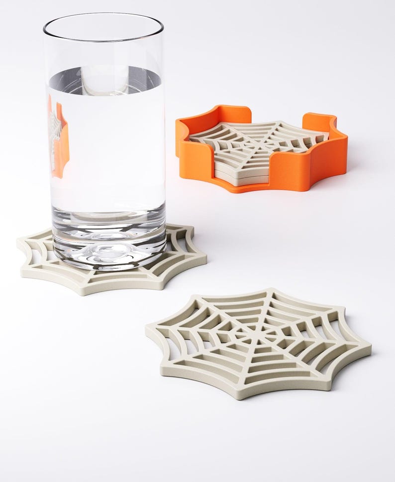 May include: A set of spiderweb-shaped coasters in light gray, with an orange holder. A tall glass of water sits on one coaster. The coasters are made of a durable material and are designed to protect surfaces from water rings. The set is ideal for Halloween.