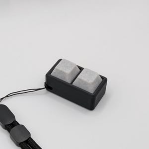 May include: A black rectangular device with two gray, speckled square buttons. The device is attached to a black lanyard with a loop and a clasp. The background is plain white.
