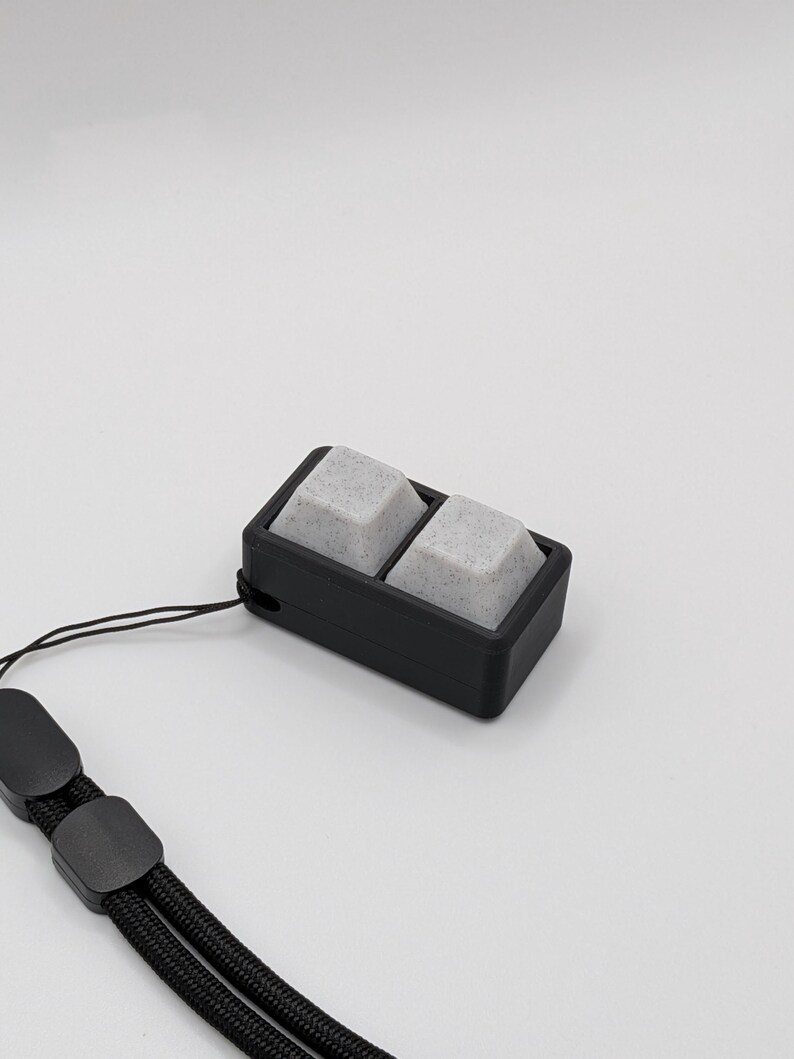 May include: A black rectangular device with two gray, speckled square buttons. The device is attached to a black lanyard with a loop and a clasp. The background is plain white.