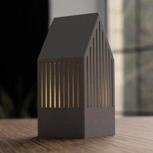 May include: A dark gray, house-shaped decorative lantern with vertical gold-colored slits. The lantern sits on a wooden surface, with a blurred background of a window and dark wall.