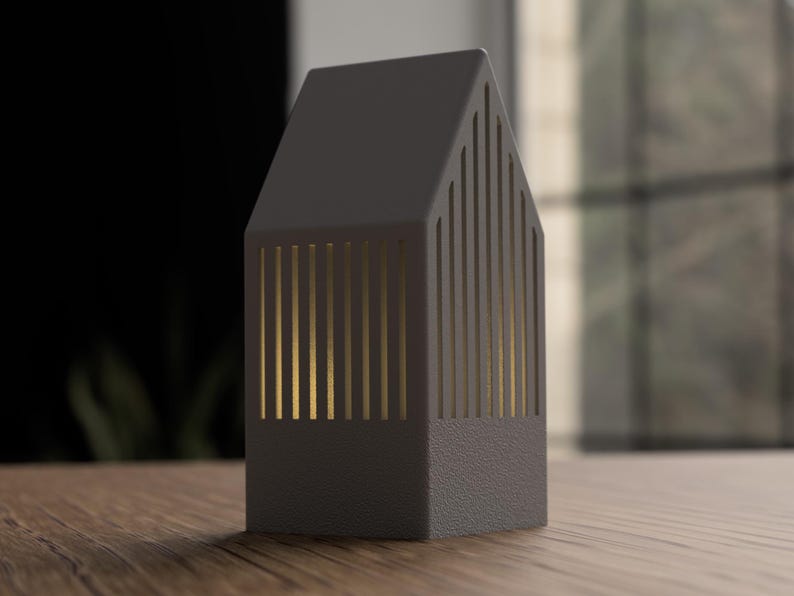 May include: A dark gray, house-shaped decorative lantern with vertical gold-colored slits. The lantern sits on a wooden surface, with a blurred background of a window and dark wall.
