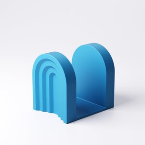 May include: A vibrant blue bookend with an arch design. One side features a layered, curved design, while the other is a solid arch. The bookend is made of a solid material and sits on a white surface.