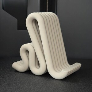 Puede incluir: Un soporte para tel&eacute;fono impreso en 3D, de color beige claro y de forma abstracta. El soporte tiene un dise&ntilde;o curvo en forma de ola con ranuras verticales en la parte posterior. El soporte est&aacute; sobre una superficie gris oscuro.
