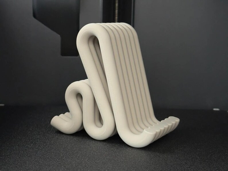 Puede incluir: Un soporte para tel&eacute;fono impreso en 3D, de color beige claro y de forma abstracta. El soporte tiene un dise&ntilde;o curvo en forma de ola con ranuras verticales en la parte posterior. El soporte est&aacute; sobre una superficie gris oscuro.