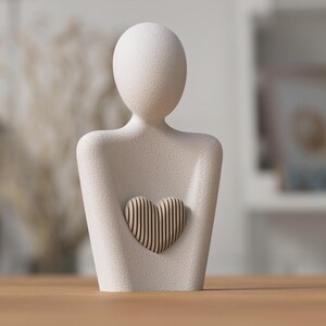 Puede incluir: Una escultura de color crema de una persona sosteniendo un coraz&oacute;n. La figura tiene una superficie lisa y texturizada. El coraz&oacute;n est&aacute; hecho de listones de madera verticales. La escultura es un objeto decorativo.
