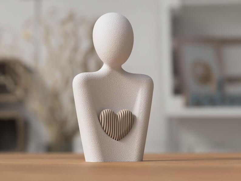 Puede incluir: Una escultura de color crema de una persona sosteniendo un coraz&oacute;n. La figura tiene una superficie lisa y texturizada. El coraz&oacute;n est&aacute; hecho de listones de madera verticales. La escultura es un objeto decorativo.