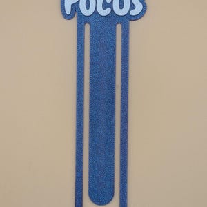 May include: A blue glittery bookmark shaped like a large paperclip with the word Focus written in white letters at the top. The bookmark has a long, slender, U-shaped design with a textured, sparkling finish throughout the entire blue surface.