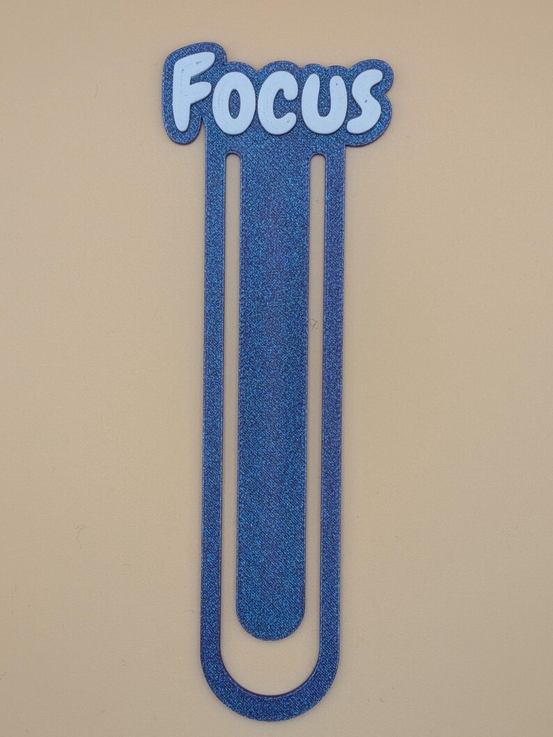 May include: A blue glittery bookmark shaped like a large paperclip with the word Focus written in white letters at the top. The bookmark has a long, slender, U-shaped design with a textured, sparkling finish throughout the entire blue surface.