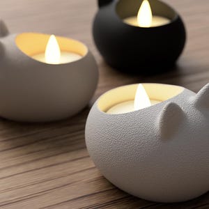 May include: Three cat-shaped candle holders in white and black, each with a lit, warm-toned LED candle. The holders are on a wooden surface, creating a cozy and decorative ambiance. The cat ear design adds a playful touch to the home decor.