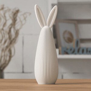 May include: A cream-colored, vertical bunny figurine with long ears. The body is a ribbed, oval shape. The figurine is on a wooden surface, with a blurred background of decorative items.