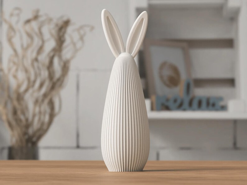 May include: A cream-colored, vertical bunny figurine with long ears. The body is a ribbed, oval shape. The figurine is on a wooden surface, with a blurred background of decorative items.