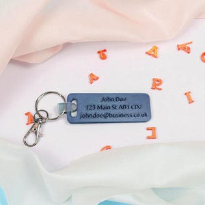 May include: A blue rectangular keychain with a silver key ring and clasp. The keychain has the name "John Doe", an address, and an email address printed on it. Orange letter-shaped confetti is scattered around the keychain.