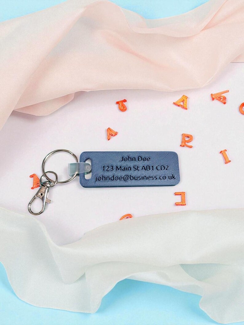 May include: A blue rectangular keychain with a silver key ring and clasp. The keychain has the name "John Doe", an address, and an email address printed on it. Orange letter-shaped confetti is scattered around the keychain.