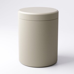 May include: A cylindrical container with a lid. The container is a light beige color and features vertical ribbed detailing on the exterior. The lid is a flat, circular shape, matching the container's color. The design is minimalist and modern.