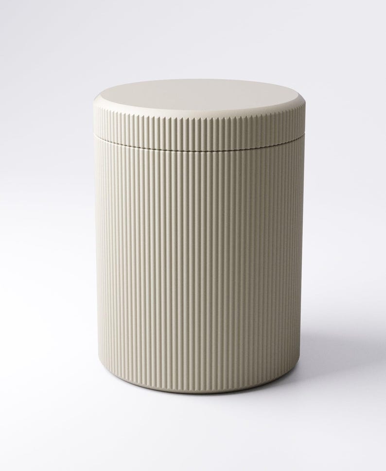 May include: A cylindrical container with a lid. The container is a light beige color and features vertical ribbed detailing on the exterior. The lid is a flat, circular shape, matching the container's color. The design is minimalist and modern.