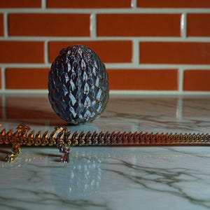 Op de afbeelding: Een gedetailleerd, metalen drakenei en een bijpassend drakenbeeldje worden getoond op een marmeren oppervlak. Het ei is een donker, getextureerd ovaal, en de draak is een lange, gelede figuur met een stekelige ruggengraat.