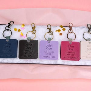 May include: A collection of personalized keychains in various colors, including navy, black, lavender, magenta, and cream. Each square keychain features the name "John Doe", an address, and a phone number, with a metal ring and clasp.