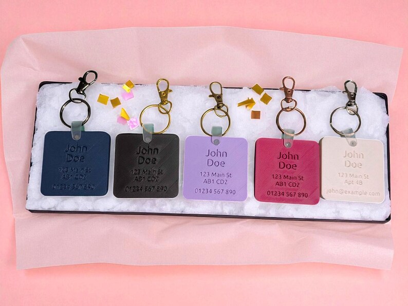 May include: A collection of personalized keychains in various colors, including navy, black, lavender, magenta, and cream. Each square keychain features the name "John Doe", an address, and a phone number, with a metal ring and clasp.