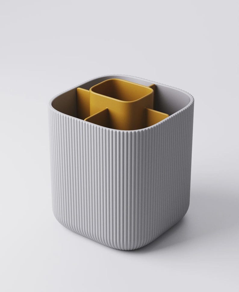 May include: A grey, ribbed desk tidy with a mustard yellow interior. The organiser has a square shape with rounded corners and vertical lines on the exterior. The interior is divided into three compartments.