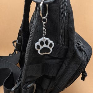 May include: A black tactical backpack with a silver key ring and a paw print charm. The paw print charm is white with a black paw print in the center. The backpack has multiple pockets and straps.