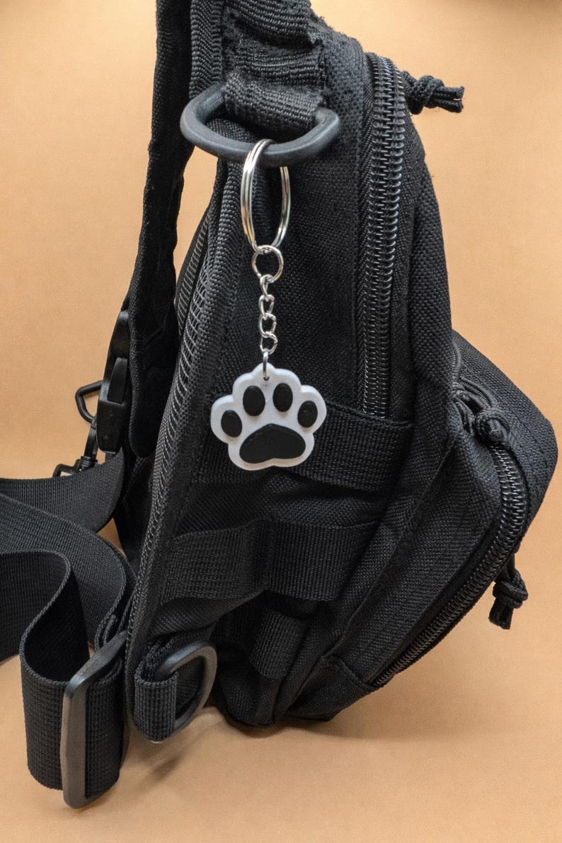 May include: A black tactical backpack with a silver key ring and a paw print charm. The paw print charm is white with a black paw print in the center. The backpack has multiple pockets and straps.