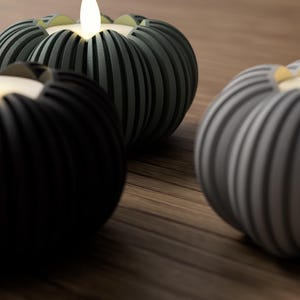May include: Three decorative candle holders with lit, flickering LED candles. The holders are pumpkin-shaped with vertical ribbed detailing. One is black, one is gray, and the other is a dark green color. The holders sit on a wooden surface.