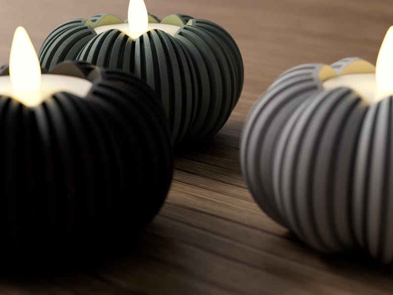 May include: Three decorative candle holders with lit, flickering LED candles. The holders are pumpkin-shaped with vertical ribbed detailing. One is black, one is gray, and the other is a dark green color. The holders sit on a wooden surface.