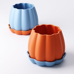 May include: Two ceramic pumpkin-shaped planters. One is light blue, the other is orange. Each planter sits on a matching scalloped-edge saucer. The planters have a ribbed texture and a wavy top edge.