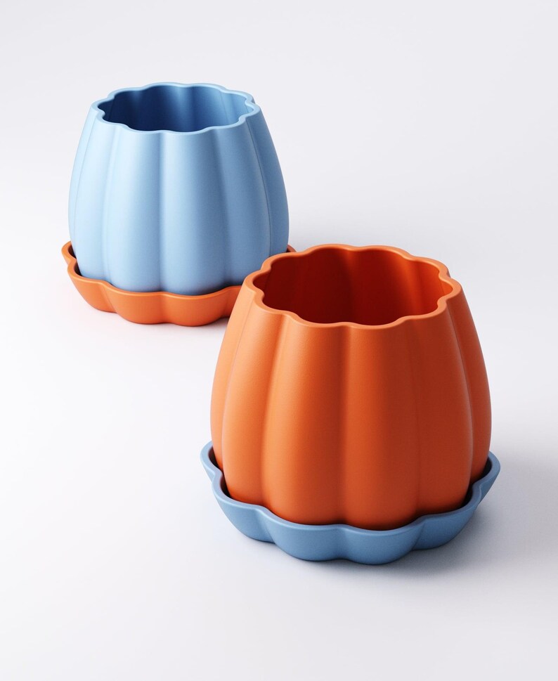 May include: Two ceramic pumpkin-shaped planters. One is light blue, the other is orange. Each planter sits on a matching scalloped-edge saucer. The planters have a ribbed texture and a wavy top edge.