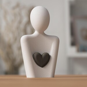 Puede incluir: Una escultura texturizada de color blanquecino de una persona sosteniendo un coraz&oacute;n. El coraz&oacute;n est&aacute; hecho de madera oscura con rayas verticales. La escultura est&aacute; sobre una superficie de madera. El fondo est&aacute; borroso, con indicios de otros objetos.