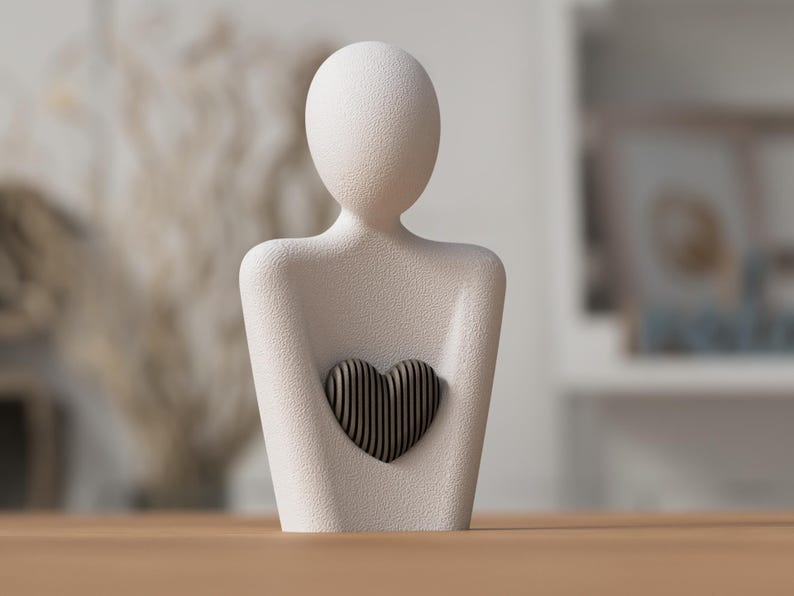 Puede incluir: Una escultura texturizada de color blanquecino de una persona sosteniendo un coraz&oacute;n. El coraz&oacute;n est&aacute; hecho de madera oscura con rayas verticales. La escultura est&aacute; sobre una superficie de madera. El fondo est&aacute; borroso, con indicios de otros objetos.