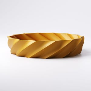 May include: Mustard yellow decorative bowl with a spiral design. The bowl has a wide, oval shape and a textured surface. The sides of the bowl are angled, creating a geometric pattern. The bowl is made of a solid material and sits on a white background.