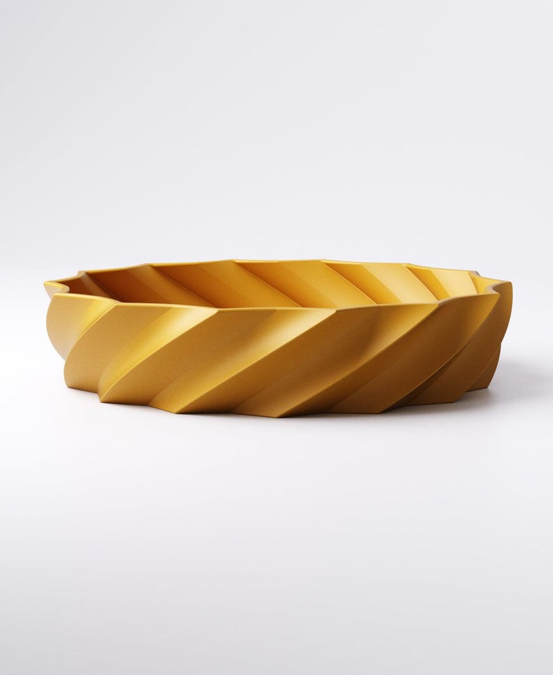 May include: Mustard yellow decorative bowl with a spiral design. The bowl has a wide, oval shape and a textured surface. The sides of the bowl are angled, creating a geometric pattern. The bowl is made of a solid material and sits on a white background.