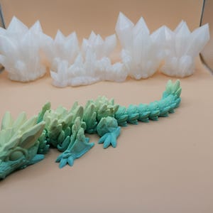 K&ouml;nnte beinhalten: Ein 3D-gedruckter, beweglicher Drache in Gr&uuml;n- und Blaut&ouml;nen mit kristallartiger Textur. Im Hintergrund befinden sich mehrere wei&szlig;e Kristallformationen. Der Drache hat einen detaillierten Kopf, Beine und einen segmentierten K&ouml;rper.