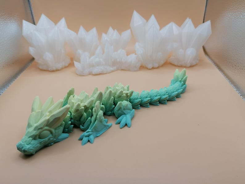 K&ouml;nnte beinhalten: Ein 3D-gedruckter, beweglicher Drache in Gr&uuml;n- und Blaut&ouml;nen mit kristallartiger Textur. Im Hintergrund befinden sich mehrere wei&szlig;e Kristallformationen. Der Drache hat einen detaillierten Kopf, Beine und einen segmentierten K&ouml;rper.
