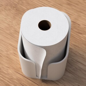 Op de afbeelding: Een witte papieren rol zit in een lichtgrijze houder met verticale ribbels. De houder staat op een houten oppervlak. De papieren rol heeft een donker centraal gat en een deel van het papier is afgerold.