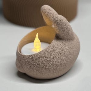 K&ouml;nnte beinhalten: Ein beigefarbener Kerzenhalter aus Keramik in Form einer Hand mit strukturierter Oberfl&auml;che. Der Kerzenhalter enth&auml;lt eine kleine, leuchtende LED-Kerze. Der Hintergrund zeigt ein verschwommenes, zylindrisches Objekt.