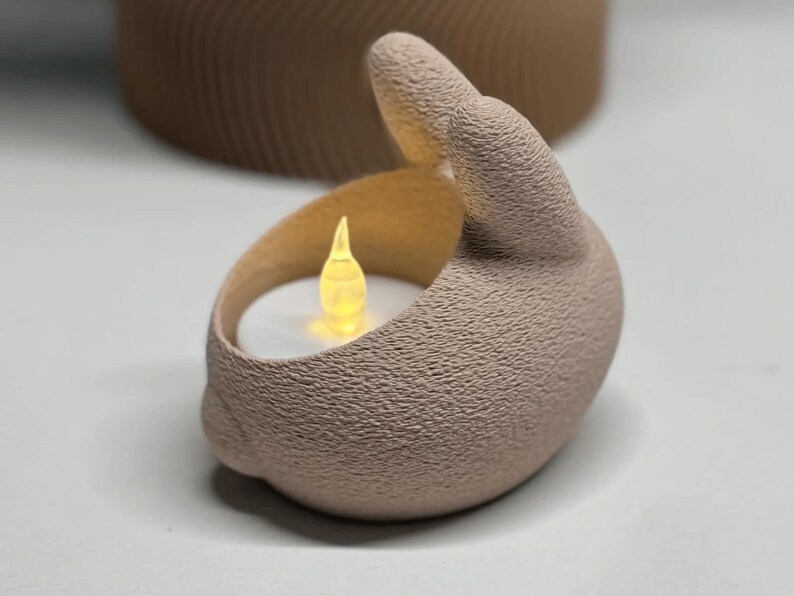 K&ouml;nnte beinhalten: Ein beigefarbener Kerzenhalter aus Keramik in Form einer Hand mit strukturierter Oberfl&auml;che. Der Kerzenhalter enth&auml;lt eine kleine, leuchtende LED-Kerze. Der Hintergrund zeigt ein verschwommenes, zylindrisches Objekt.