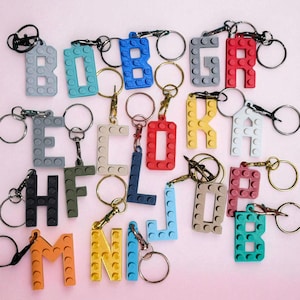 May include: A collection of colorful letter keychains, each constructed from interlocking building blocks. The keychains feature various letters in shades of gray, blue, red, yellow, and orange, with silver key rings attached. The background is a soft pink.