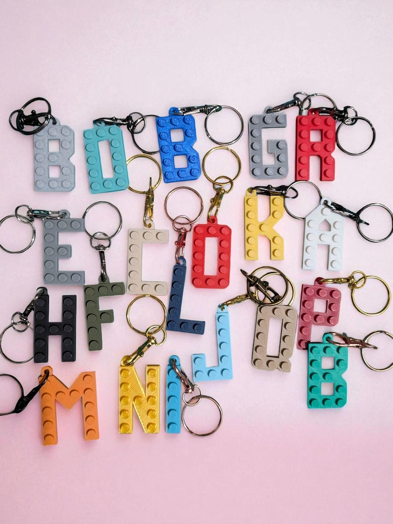 May include: A collection of colorful letter keychains, each constructed from interlocking building blocks. The keychains feature various letters in shades of gray, blue, red, yellow, and orange, with silver key rings attached. The background is a soft pink.
