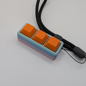 May include: A custom mechanical keyboard with three orange keycaps. The keycaps are set in a gradient case of blue, pink, and purple. A black lanyard is attached to the keyboard.