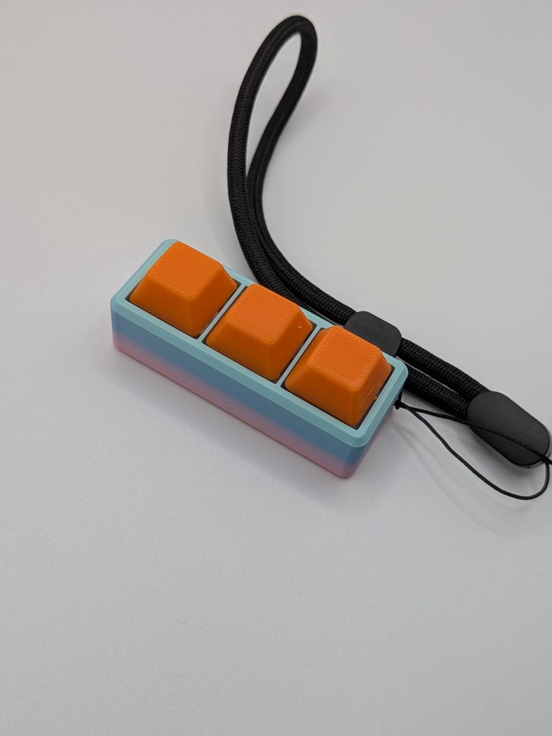 May include: A custom mechanical keyboard with three orange keycaps. The keycaps are set in a gradient case of blue, pink, and purple. A black lanyard is attached to the keyboard.