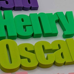 May include: Green and yellow 3D printed name blocks spelling out "Henry" and "Oscar". The letters are thick and blocky, with a slightly rounded font. Other name blocks in purple and pink are partially visible in the background.