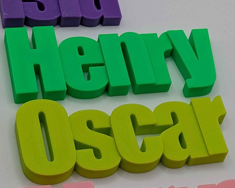 May include: Green and yellow 3D printed name blocks spelling out "Henry" and "Oscar". The letters are thick and blocky, with a slightly rounded font. Other name blocks in purple and pink are partially visible in the background.